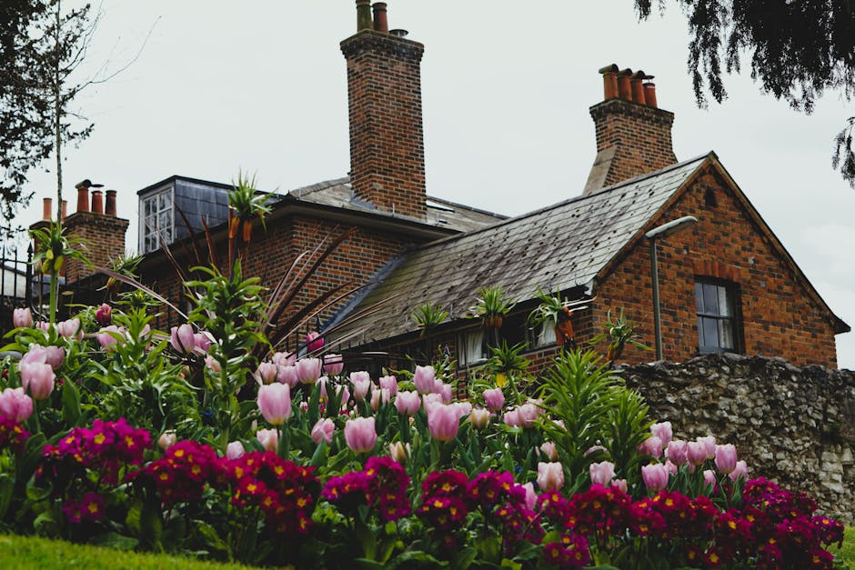 A traditional brick house with a pitched slate roof and multiple chimneys is surrounded by a lush garden. In the foreground, there is a vibrant flower bed filled with pink tulips and deep red primrose